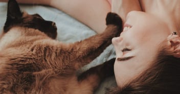 Woman with brown hair lying down looking happy with Siamese cat
